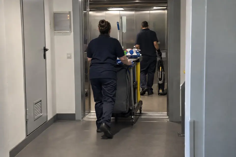 Dos trabajadores de limpieza de espaldas entrando en un montacargas con un carro y un aspirador por un pasillo de servicio