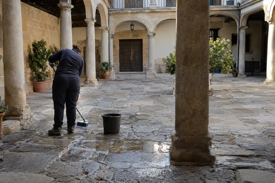 Operaria frotando con un cepillo el enlosado de piedra del patio porticado de un edificio histórico, con un cubo al lado