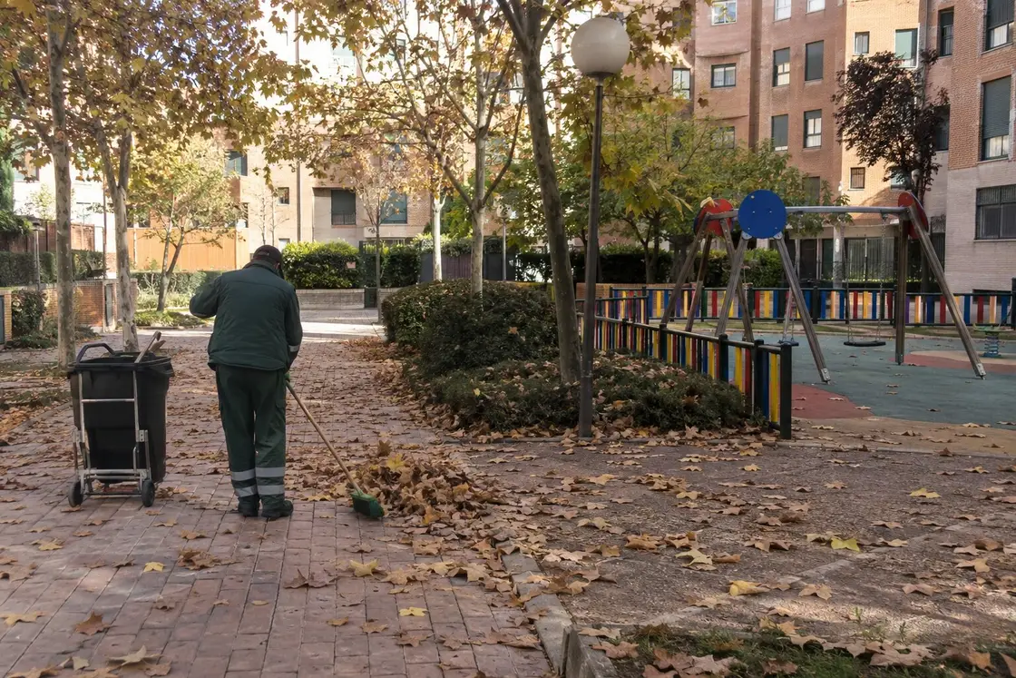 Operario barriendo hojas secas en el paseo de una urbanización, junto a un parque infantil y un contenedor con ruedas