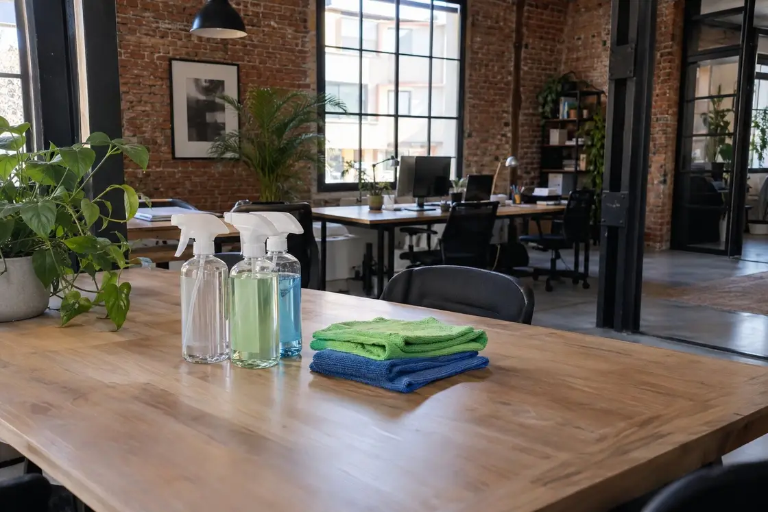 Pulverizadores rellenables y bayetas de microfibra verdes y azules sobre una mesa de madera en una oficina tipo loft