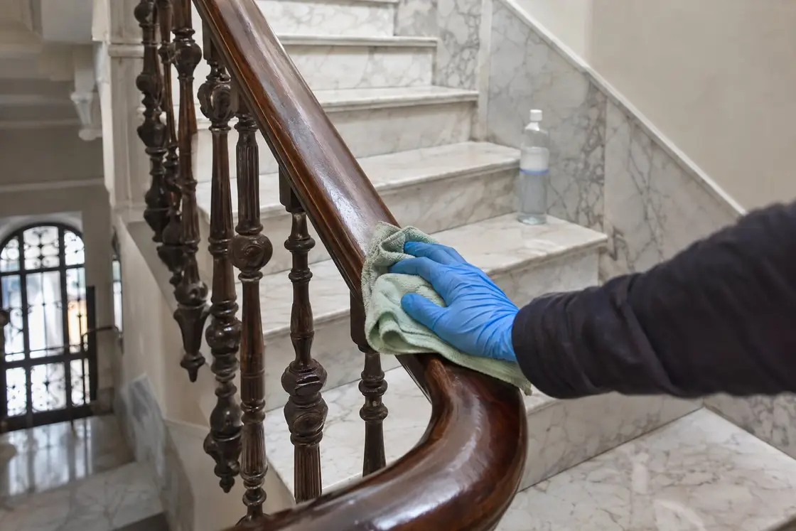 Mano con guante de nitrilo limpiando con una bayeta el pasamanos de madera de una escalera de mármol blanco