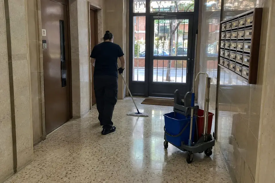 Limpiadora de espaldas fregando con mopa el terrazo del portal de una comunidad, junto a un carro de dos cubos y los buzones