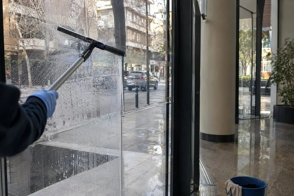 Mano con guante azul pasando una rasqueta por la cristalera de un vestíbulo de oficinas, con la calle al otro lado