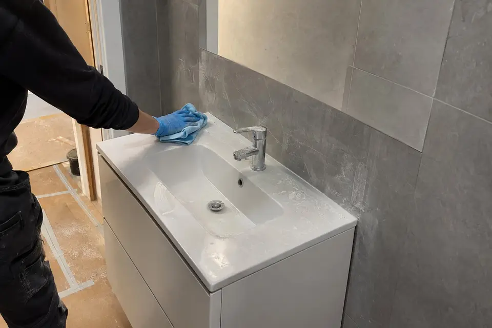 Mano con guante quitando con una bayeta el polvo de obra de un lavabo nuevo en un baño recién reformado con azulejo gris