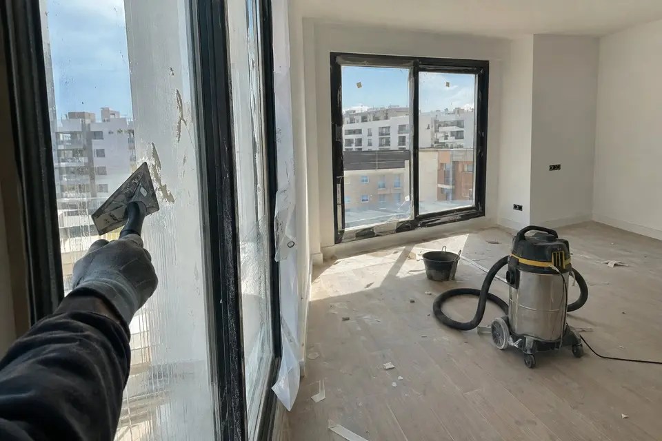 Mano con guante rascando restos de pintura del cristal de una ventana en un piso recién terminado, con un aspirador de obra