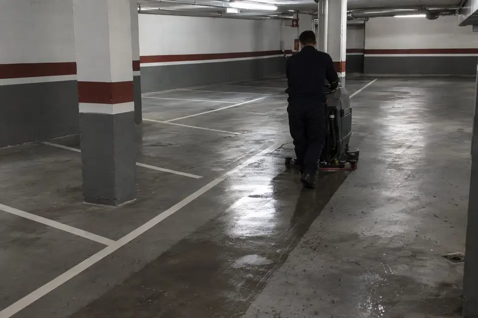 Operario de espaldas con una fregadora que deja una franja limpia y húmeda en el suelo de un garaje comunitario vacío