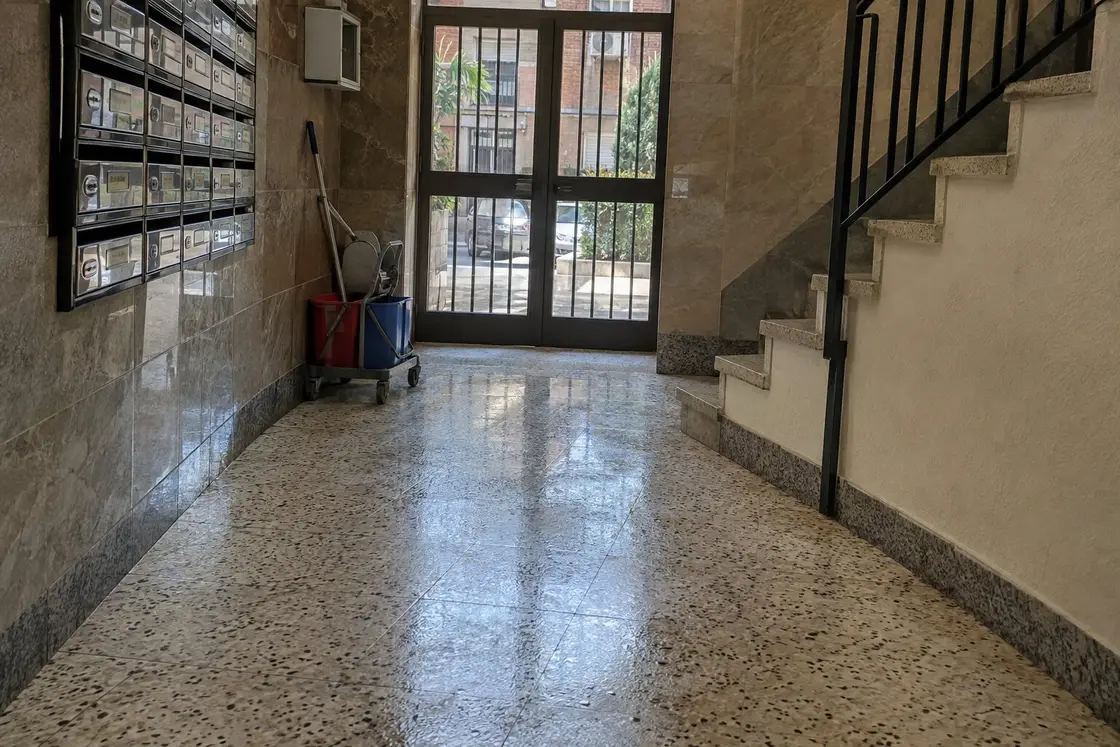 Portal de un bloque de los años 70 con el terrazo recién fregado, buzones metálicos y un carro de fregado de dos cubos