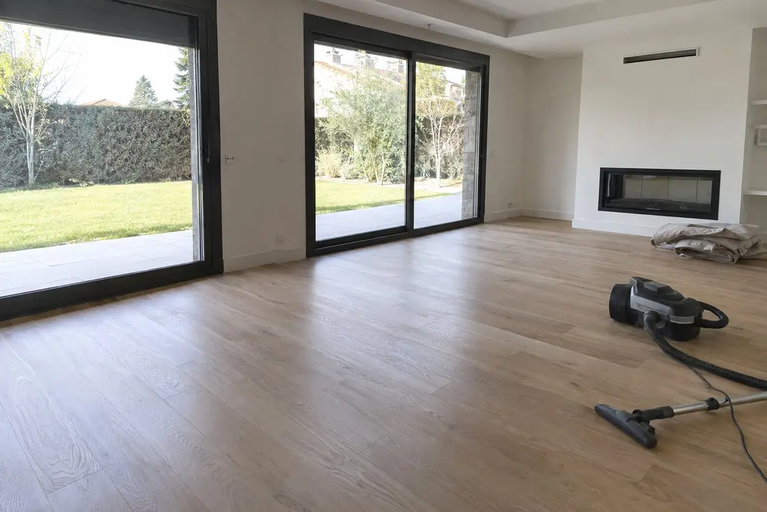 Salón vacío de un chalet tras una reforma, con el parquet de roble claro limpio, ventanales al jardín y un aspirador