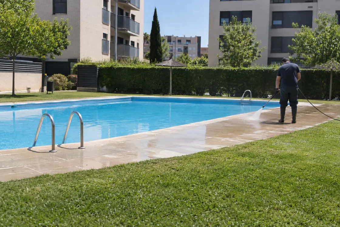 Operario limpiando con hidrolimpiadora el borde de piedra de una piscina comunitaria, con césped y bloques de pisos