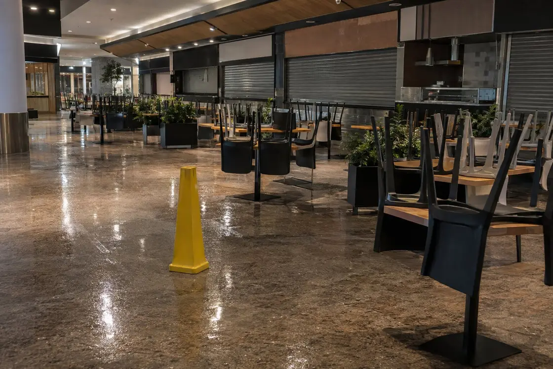Zona de restauración de un centro comercial ya cerrada, con las sillas sobre las mesas, el suelo fregado y un cono amarillo